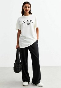 Witte T-shirt met de tekst "PILATES Club", gecombineerd met zwarte wijde broek en een gequilt zwarte tas. Glad stof, ontspannen pasvorm.