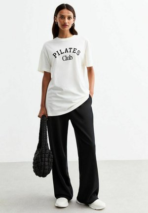 Witte T-shirt met de tekst "PILATES Club", gecombineerd met zwarte wijde broek en een gequilt zwarte tas. Glad stof, ontspannen pasvorm.
