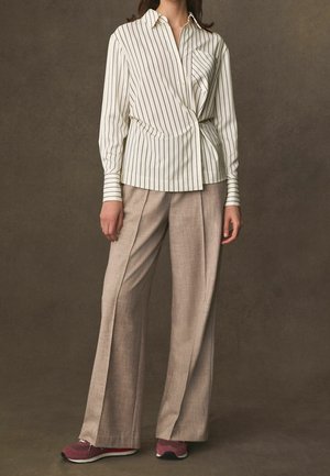 Vrouw draagt een wit overhemd met lange mouwen en spijkerpatroon, beige broek met wijde pijpen en bordeauxrode sneakers tegen een effen bruine achtergrond.