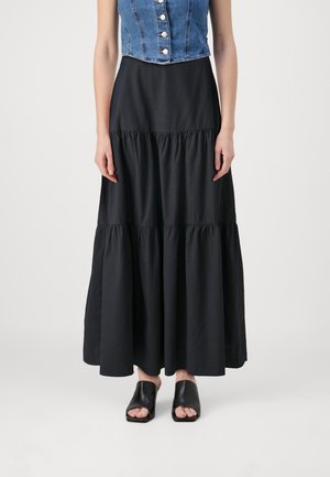 Vrouw draagt een gelaagde zwarte maxi-rok, mouwloos blauw denim gilet met knopen en zwarte open-toe schoenen met hak, gefotografeerd tegen een witte achtergrond.