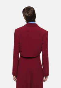 Blazer bordeaux tagliato con maniche lunghe, vestibilità aderente, collo a punta e superficie liscia, indossato sopra una camicia azzurra chiara.