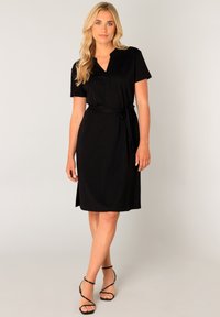 Zwart kortemouwende jurk met een V-hals en tailleband. Gladde stof met een lengte tot onder de knie en een zij-opening. Gekomplementeerd met zwarte sandalen met bandjes.