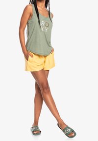 Vihreä tanktoppi, jossa on "ROXY"-logo, keltaiset sortsit sivutaskuilla ja vihreät liukukengät valkoisella brändäyksellä. Pehmeä kangas ja rento leikkaus.