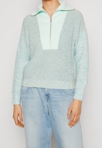 Hellblauer Strickpullover mit einem Reißverschluss am Stehkragen, der einen strukturierten Körper und eine einfarbige Vorderseite aufweist, kombiniert mit hellblauen Jeans.