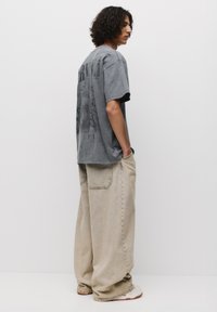 Grå oversized t-shirt med grafisk tryk på ryggen, parret med lysebeige baggy jeans. Har en afslappet pasform og ribbet halsudskæring.