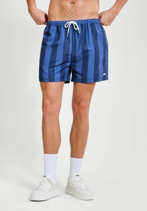Mann trägt blaue, vertikal gestreifte Badeshorts, weiße Socken und weiße Turnschuhe, steht vor einem schlichten hellen Hintergrund.