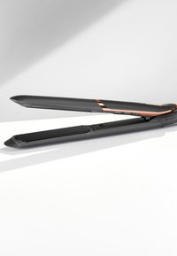 Plancha de pelo en negro y oro rosa con placas cerámicas anchas, reposando abierta sobre una superficie blanca frente a un fondo gris.