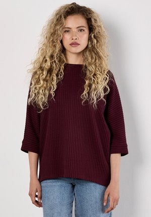 Burgundy textured top with three-quarter sleeves, relaxed fit, and crew neckline, paired with light denim jeans. Simple design, no patterns.