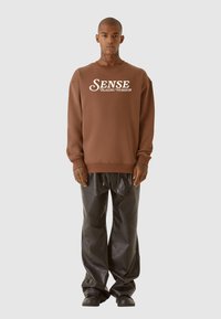 Brun sweatshirt med vit text "SENSE BLAZING HORIZON", ihopparad med glansiga svarta vida byxor och svarta skor.