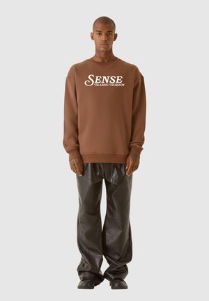 Brun sweatshirt med hvid "SENSE BLAZING HORIZON" tekst, parret med skinnende sorte brede bukser og sort fodtøj.