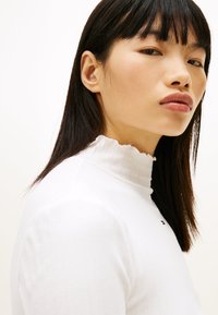 Λευκό ριμπ ζιβάγκο με λοξό γιακά και μικρή διακριτική πινελιά με λογότυπο. Ομαλή υφή και εφαρμοστό σχέδιο.