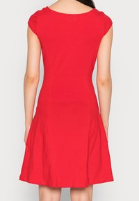 Mujer con un vestido rojo de mangas cortas y hasta la rodilla, vista desde atrás contra un fondo claro y liso.