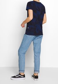 T-paita tummansinisestä kankaasta, lyhythihainen, jossa on teksturoitu abstrakti kuvio, yhdistettynä vaaleansinisiin farkkuihin ja mustiin Vans-lenkkareihin.