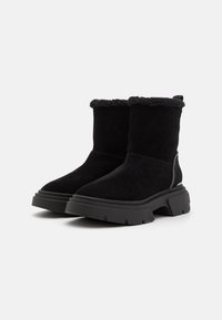 Bottines en suede noir avec une épaisse semelle noire, un col duveteux et des accents sur les côtés. Texture lisse avec un design à bout arrondi.