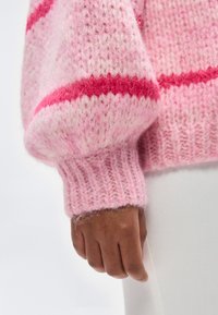 Χέρι με σκούρο δέρμα που φοράει ρόζ χνουδωτό πλεκτό πουλόβερ με μια κόκκινη οριζόντια ρίγα, συνδυασμένο με λευκό παντελόνι πάνω σε απλό φόντο.