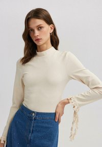 Beige ribbeltrui met lange mouwen en hoge halslijn, versierd met crèmekleurige franjes op de mouwen, gecombineerd met een blauwe denim rok met versieringen.