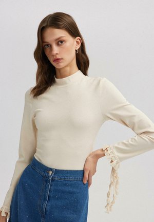 Haut beige à manches longues côtelé avec un col montant et des accents à franges crème sur les manches, associé à une jupe en denim bleu ornée de décorations.