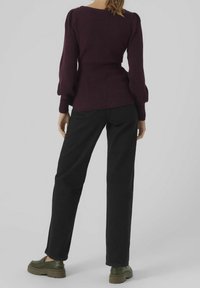 Eine Frau steht mit dem Rücken zur Kamera und trägt einen dunkelvioletten Pullover, schwarze gerade Hosen und olivgrüne klobige Schuhe.