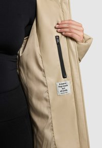 Chaqueta acolchada color beige con una textura suave, que cuenta con un bolsillo interior con cremallera y una etiqueta que dice: "Un abrazo cálido hecho de tela."
