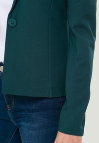 Zelený přiléhavý blejzr s hladkou texturou, zapínání na jeden knoflík a dvěma bočními kapsami, kombinovaný s modrými džínami.