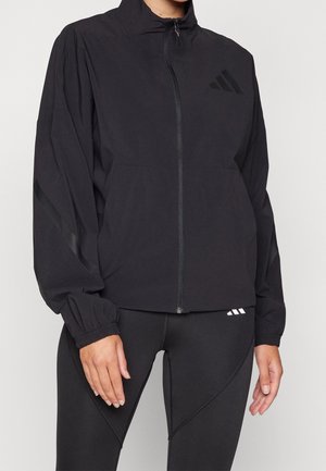 Personne portant une veste zippée noire avec des poignets élastiques et un legging noir assorti, tous deux arborant des logos Adidas discrets.