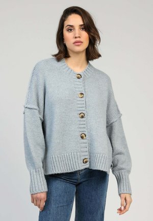 Jeune femme aux cheveux bruns mi-longs portant un pull en grosse maille bleu pâle boutonné et un jean bleu sur un fond uni.