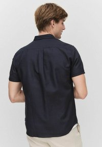 Kortärmad marinblå skjorta i lätt tyg, med spetsig krage, bakfållar och rundad nederkant, som vilar på off-white byxor.