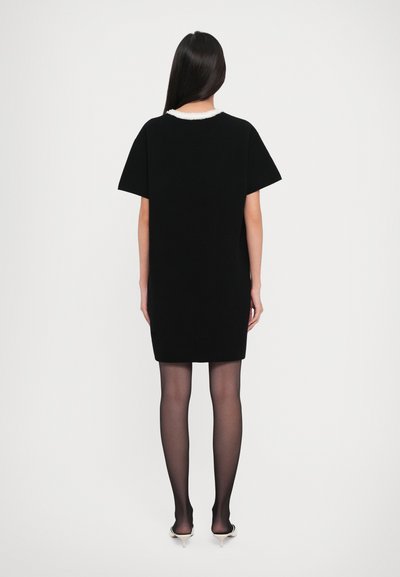 Woman with long dark hair wears a black short-sleeve dress with white collar, sheer black tights, and white high-heeled shoes, standing facing away.