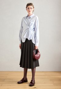 Chemise bleu clair avec un devant torsadé, jupe plissée grise, sac en cuir bordeaux, collants noirs et mocassins avec un accent doré.