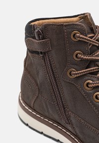 Close-up van een bruine leren laars met tan en bruine veters, een zijrits, metalen oogjes, een gevoerde kraag en een witte gestikte zool.