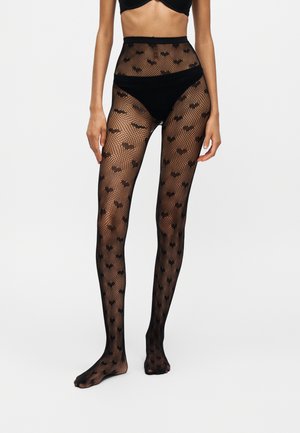 Femme portant des collants en résille noirs avec un motif de chauve-souris, assortis à des sous-vêtements noirs et un soutien-gorge noir, debout sur un fond uni.
