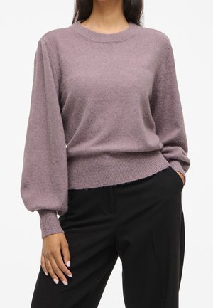 Kvinde iført en mauve langærmet striktrøje med ballonermer og sorte bukser, den ene hånd i lommen og den anden hvilende ved siden.