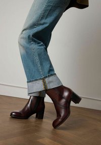Legs wearing cuffed blue jeans and brown leather ankle boots with block heels on a wooden floor against a plain wall.