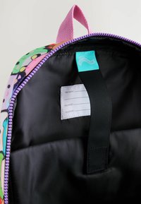 Lila und pink gemusterter Rucksack-Innenraum mit einem schwarzen Futter, ausgestattet mit einem Namensschildhalter und einem breiten Reißverschluss. Weiche Textur, strapazierfähiges Material.
