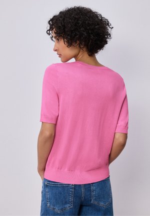 Femme aux cheveux courts et bouclés portant un pull rose à manches courtes et un jean bleu, vue de dos sur un fond clair uni.
