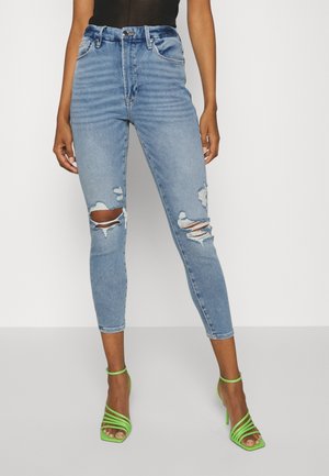 Kvinde iført lyseblå højtaljede ridsede skinny jeans og lysegrønne højhælede sandaler med stropper, står mod en hvid baggrund.