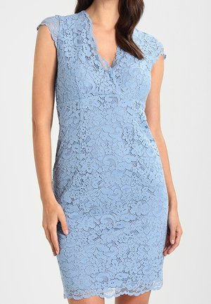Femme portant une robe en dentelle bleu clair ajustée et sans manches, avec un col en V à bords festonnés et un motif floral, sur un fond uni.