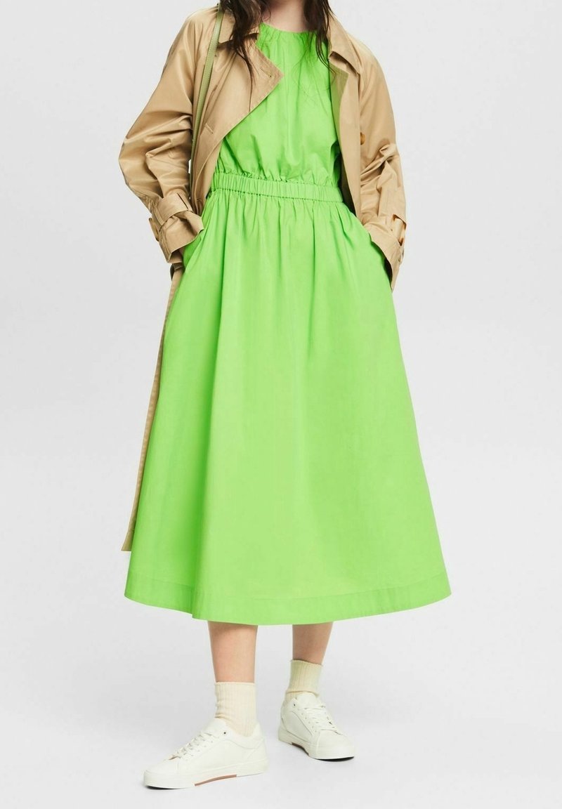Femme portant une robe midi vert vif avec taille élastique, un trench coat beige, des chaussettes blanches et des baskets blanches, posant les mains dans les poches.