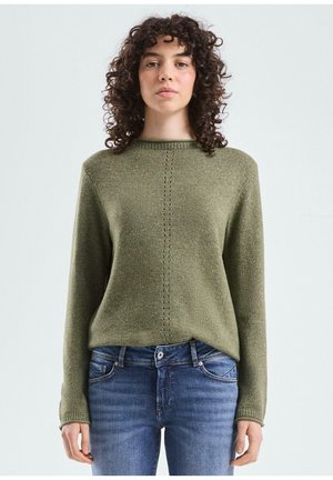 Vrouw met krullend donker haar draagt een olijfgroene gebreide trui en blauwe spijkerbroek, staand tegen een effen lichtgrijze achtergrond.