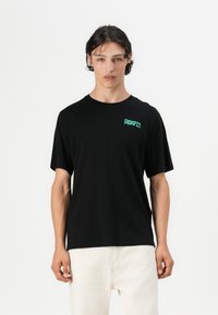 Camiseta de manga corta negra con el logo verde "ROOTS" en el lado izquierdo del pecho. Hecha de algodón, presenta un ajuste relajado y un escote redondo.