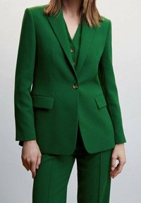 Femme portant un blazer vert ajusté avec des poches à rabat, un pantalon vert assorti et un gilet vert boutonné en dessous.