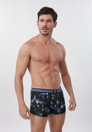 Uomo senza maglietta con capelli corti e scuri che indossa boxer neri con stampa floreale blu, in piedi davanti a uno sfondo chiaro e semplice.