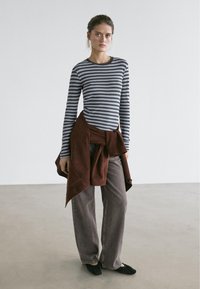 Young woman in gray and black striped long-sleeve top, loose gray pants, black shoes, with brown jacket tied around her waist, standing in empty room.