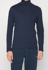 Mies, który ma na sobie granatową bluzę z długim rękawem i golfem oraz dopasowane spodnie, stoi na tle jednolitego jasnego tła.