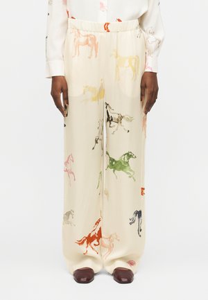 Κρεμ παντελόνια με φαρδιά μπατζάκια και πολύχρωμα σχέδια αλόγων, φορεμένα με λευκό πουκάμισο και σκούρα καφέ παπούτσια σε μοντέλο που στέκεται σε λευκό φόντο.