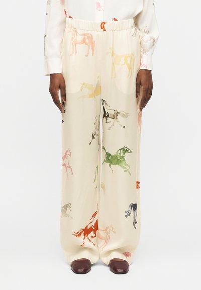 Pantalon large crème avec imprimés colorés de chevaux, porté avec une chemise blanche et des chaussures marron foncé sur un mannequin debout devant un fond blanc.