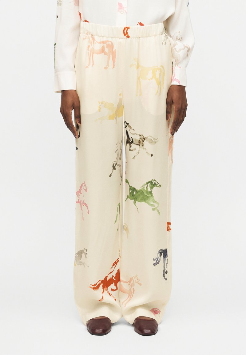 Κρεμ παντελόνια με φαρδιά μπατζάκια και πολύχρωμα σχέδια αλόγων, φορεμένα με λευκό πουκάμισο και σκούρα καφέ παπούτσια σε μοντέλο που στέκεται σε λευκό φόντο.