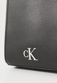 Bolso de mano de cuero negro texturizado con un logotipo plateado en relieve "cK" en la parte frontal, que destaca su diseño suave y duradero.