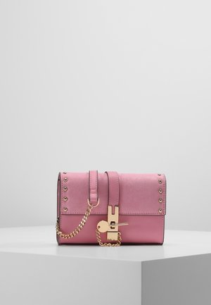 Roze leren en suède handtas met gouden studs, decoratieve gouden ketting en gouden sluiting op een wit voetstuk tegen een grijze achtergrond.