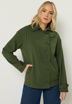 Mujer con una chaqueta verde abotonada con bolsillos, posando con una mano en el bolsillo y pantalones vaqueros azules sobre un fondo claro y liso.